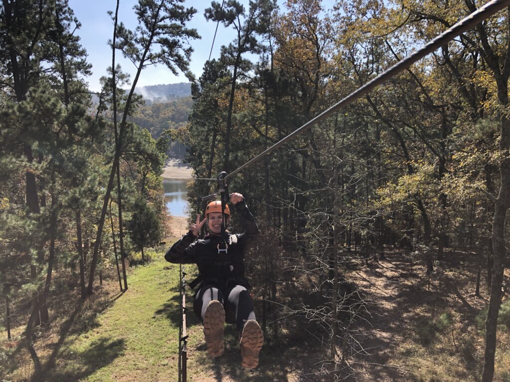 Alex Bethune zip lining surrounded by nature's scenic trees holding up a peace sign.