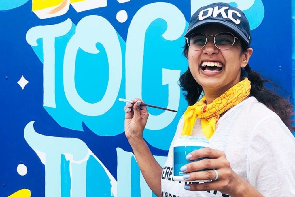 Aditi Heins smiling with a paint brush in front of a wall mural.
