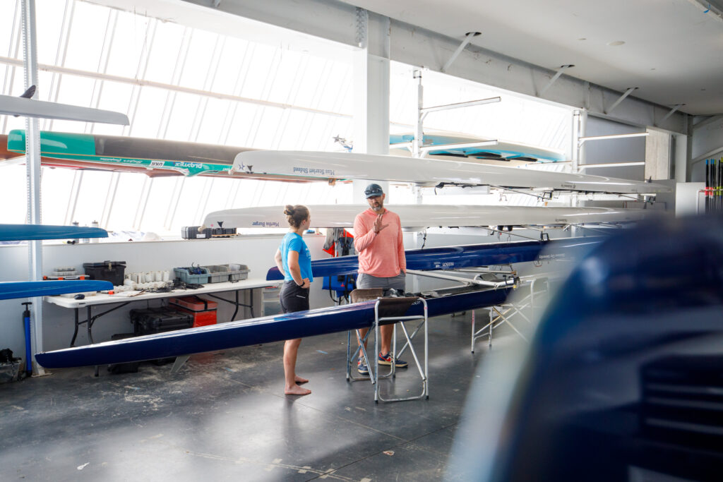 Bryan speaking with rowing athlete while standing next to various row boats inside the boathouse. 