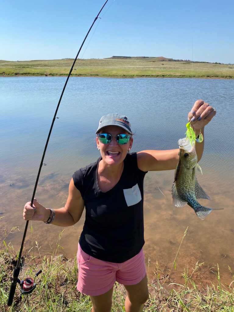 Stacey Hesser smiles big holding up a small fish she caught with a fishing rod.