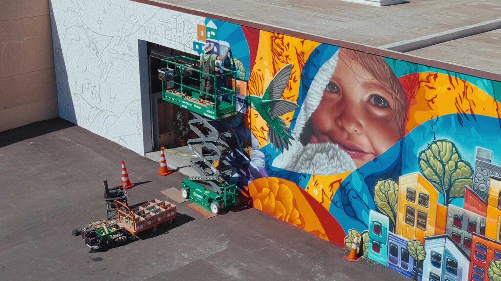 Artist and muralist Carlos Barbosa uses spray paint to create a massive, colorful public mural in Oklahoma. Here, Barbosa is shown on a scissor lift in the midst of his artistic process.